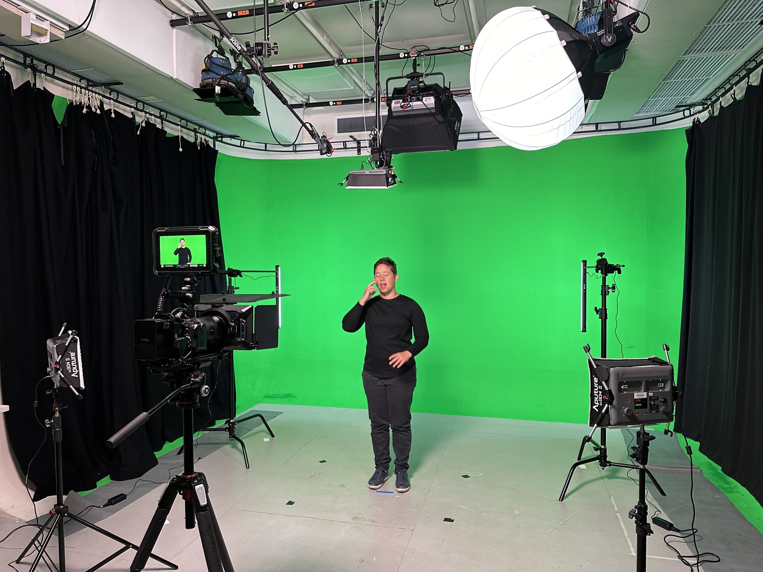 An Auslan presenter signing in a green-screen studio, surrounded by cameras and lighting.